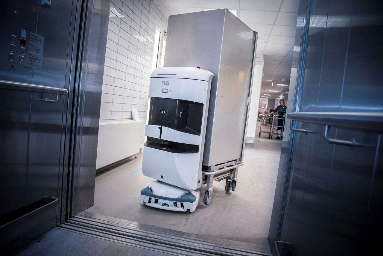 En hvid robot der kører med en hospitals madvogn. Den er på vej ind i en elevator.